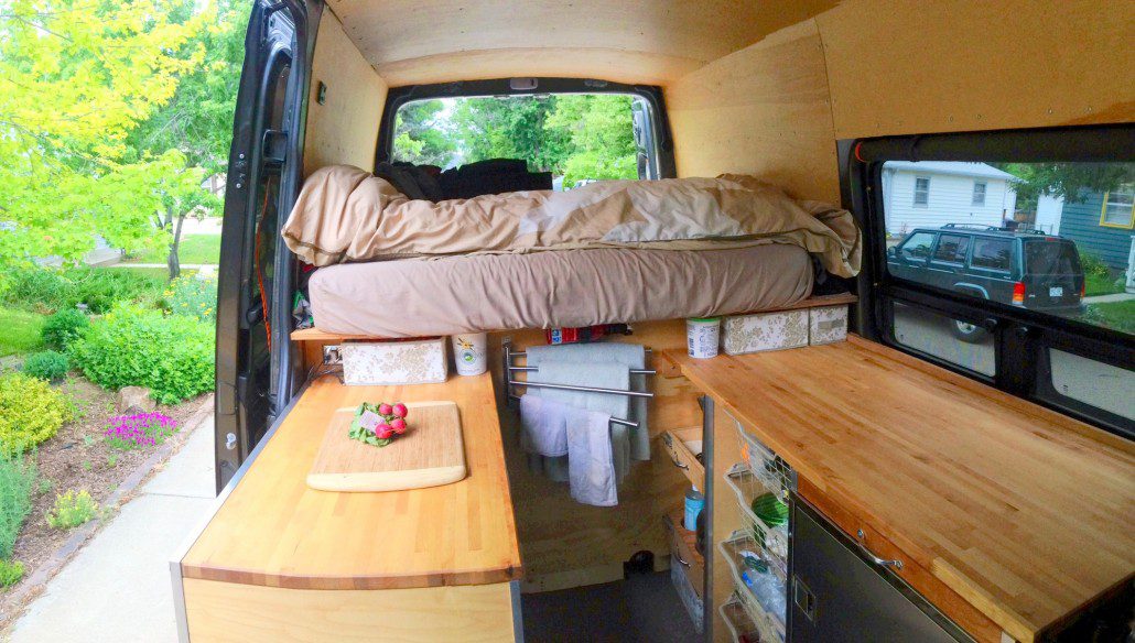 View in Sprinter van sitting in front swivel seat looking back. Ikea countertops on either side, utility drawer over fridge on drivers side, and storage with wire baskets underneath the countertops.